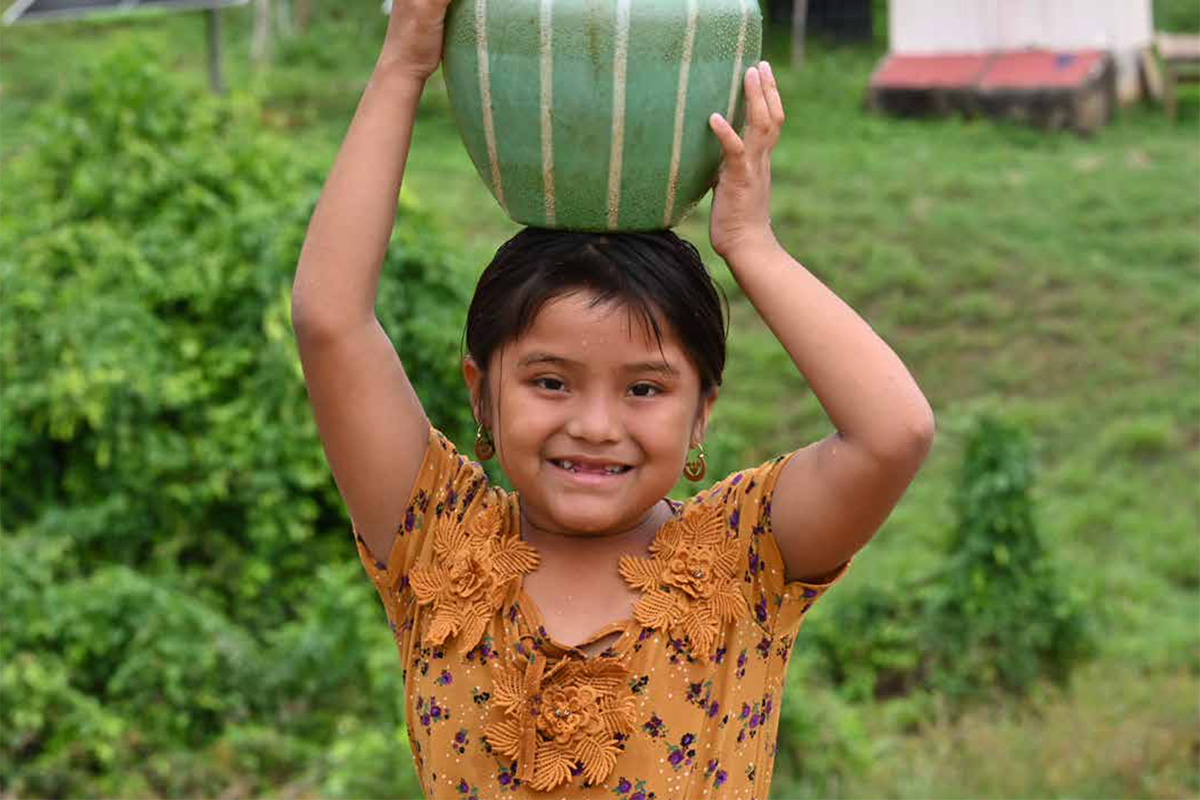 A child holding a water jug on her head stands smiling in a field